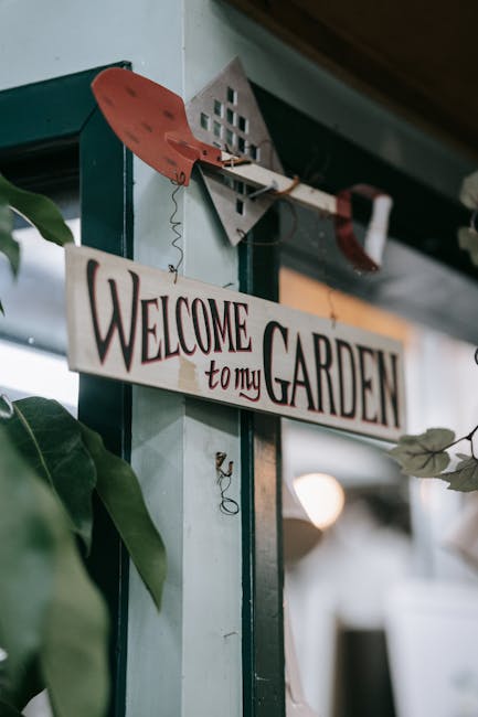 A close-up view of a garden interior featuring a wooden sign hanging from a rustic metal hook, inscribed with 'Welcome to my Garden' in decorative black and red lettering. The sign is suspended from a beam beside a green doorframe, with a leafy plant visible in the foreground. The background reveals a glimpse of the garden space, with a blurred light source and indoor elements suggesting a cozy, welcoming environment. This setting highlights the aesthetic appeal and personal touch of garden decor, emphasizing outdoor hospitality and garden maintenance themes, with no visible plants, soil, grass, or tools present in the scene. The image promotes the idea of creating inviting garden spaces, in line with the services offered by Gardeners Ealing.