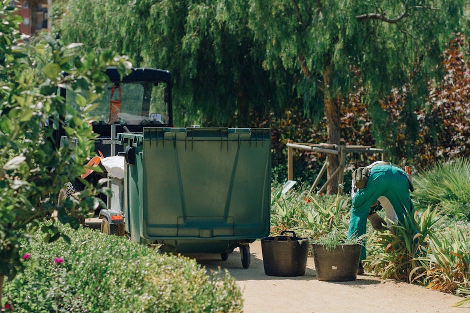 A gardener wearing a teal uniform is engaged in garden maintenance work, bending over amidst a lush garden with various plants and shrubs. The scene features a large green waste bin, and the gardener appears to be trimming or harvesting plants, with her hands close to the foliage. Nearby, there are two black baskets filled with garden clippings or tools on a paved pathway. In the background, mature trees with green and reddish leaves provide shade, and a wooden framework or trellis is visible among the dense foliage. The overall environment showcases a well-maintained outdoor space with a mix of flowering plants, leafy bushes, and neatly cut grass, under diffused natural lighting typical of a bright day, emphasizing the ongoing landscaping work by Gardeners Ealing, consistent with garden waste management and landscaping themes linked to the Ealing Council garden waste rules and gardeners compliance.